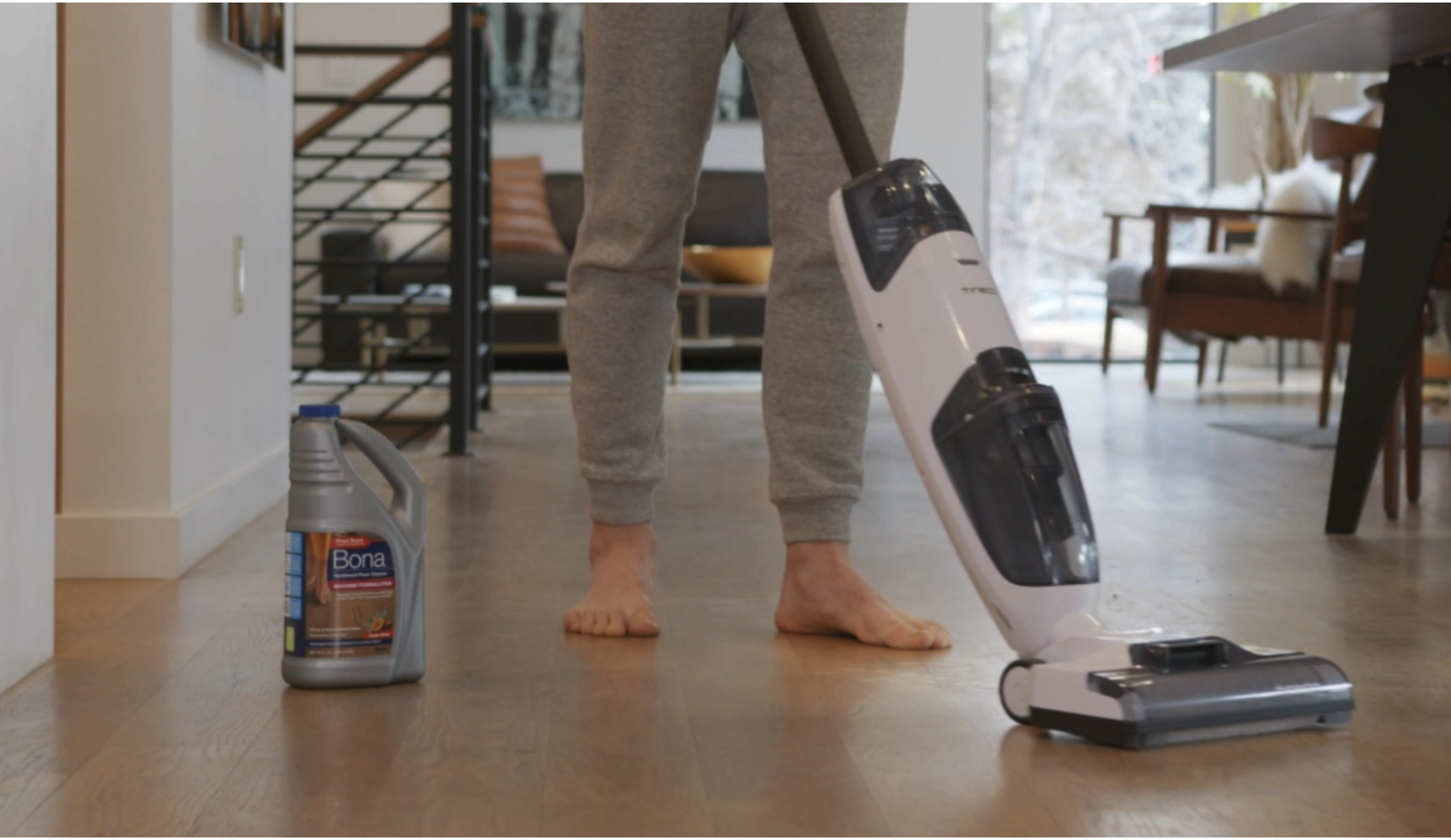A barefoot man cleans a hardwood floor with a cleaning machine and Bona Hardwood Floor Cleaner Machine Formulation.
